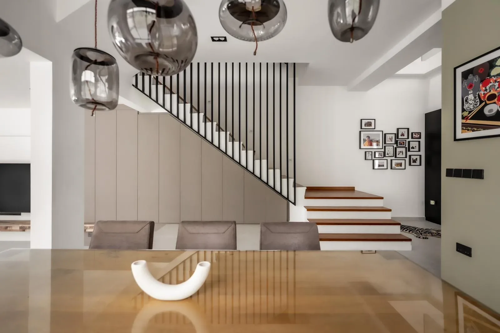 Modern dining area with brown chairs, a wooden table, and a white decorative piece. Glass light fixtures hang above like beacons in a hero story. A staircase with black railings and a photo gallery wall are in the background.